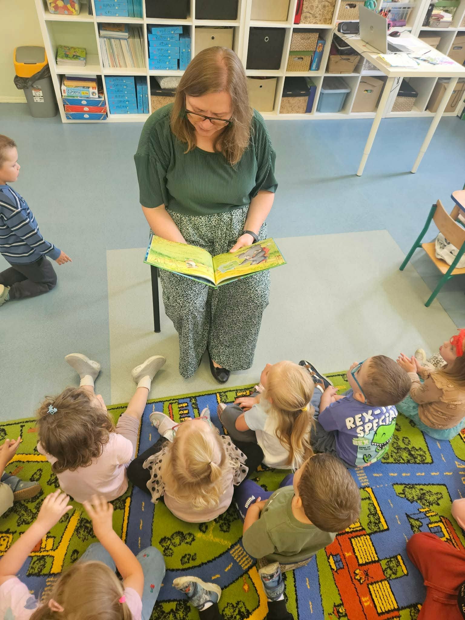 przedszkolaki siedzą na dywanie i słuchają Pani bibliotekarki, która czyta im książkę