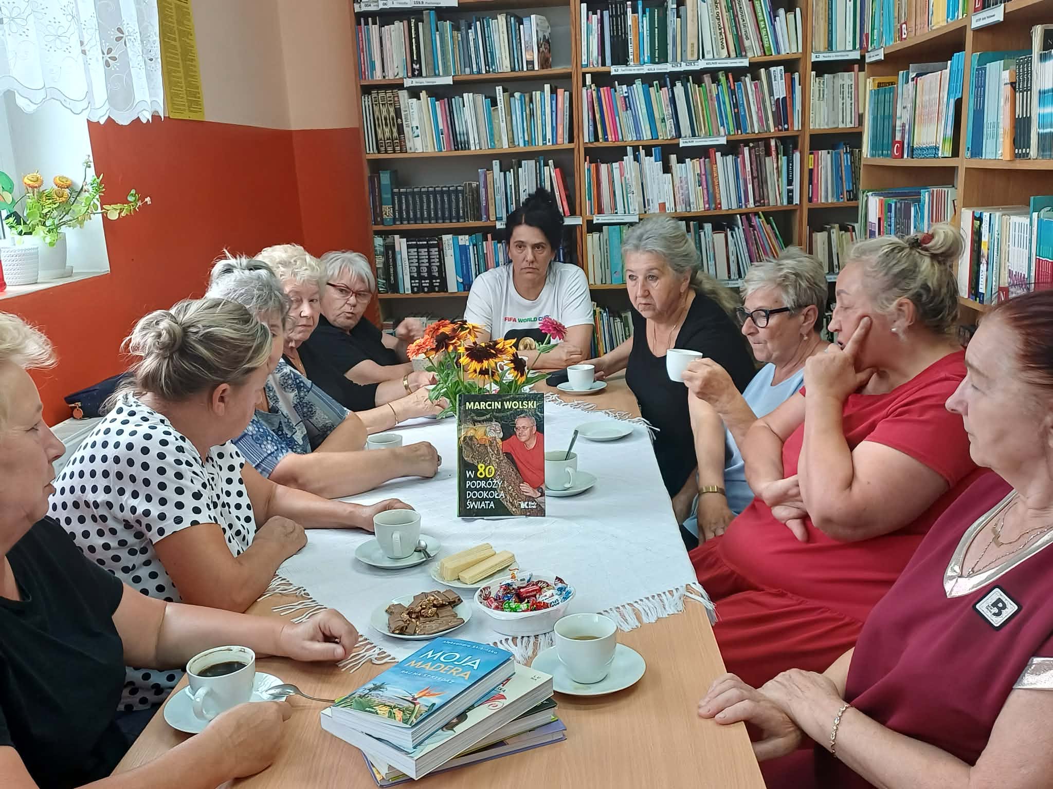 panie siedzą przy stole na którym są książki, słodycze i filiżanki