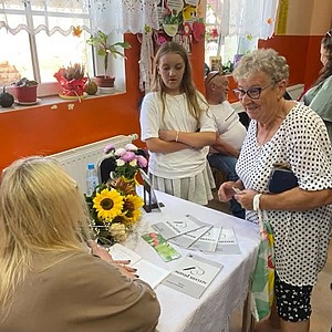 2 kobiety stoją przed siedząca na krześle autorką